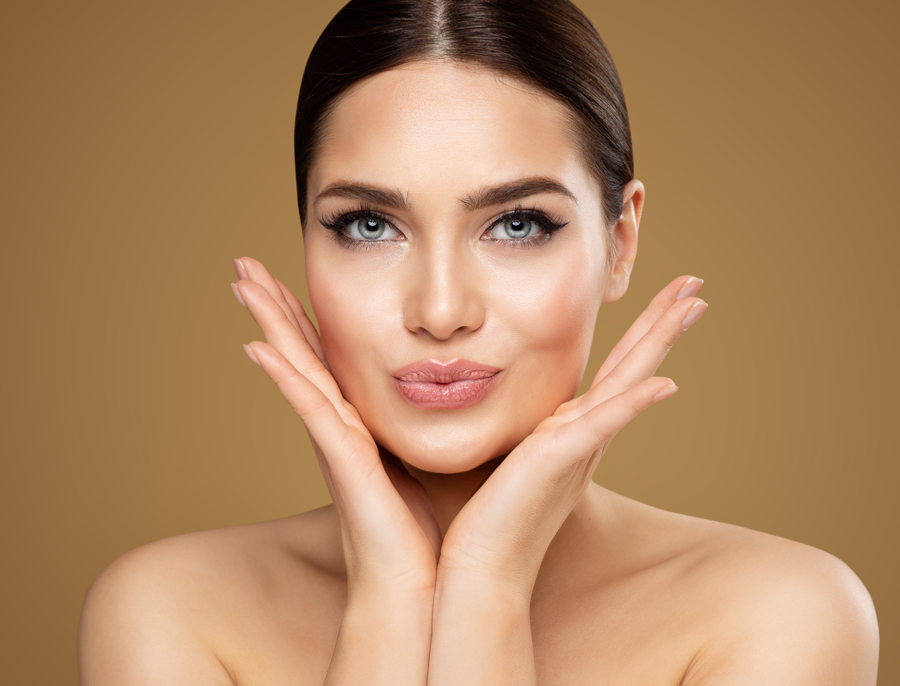 A woman with her hair pulled back rests both hands against her cheeks, photographed against a warm gold backdrop.