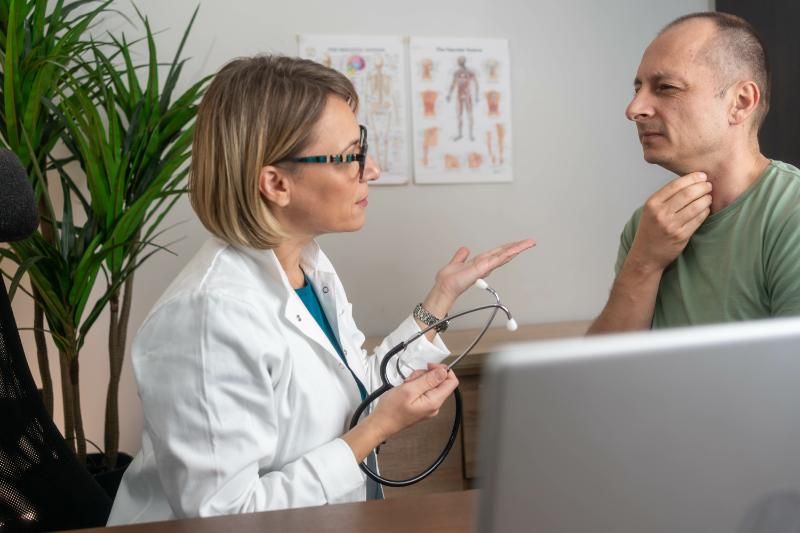A clinician in a white coat holding a stethoscope talks with a seated man in an office with an anatomy chart on the wall.