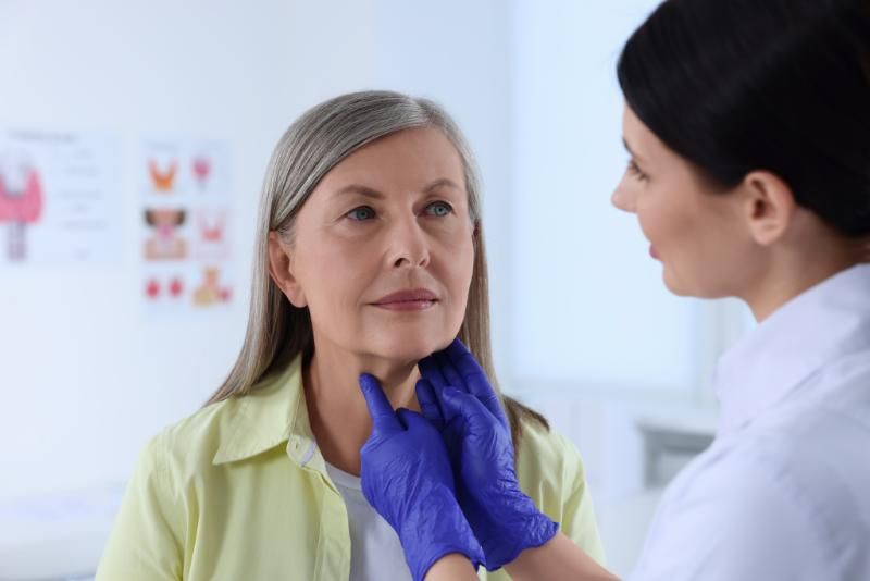 A clinician in gloves examines the neck of a seated woman with grey hair, in a clinic room with a thyroid chart on the wall.