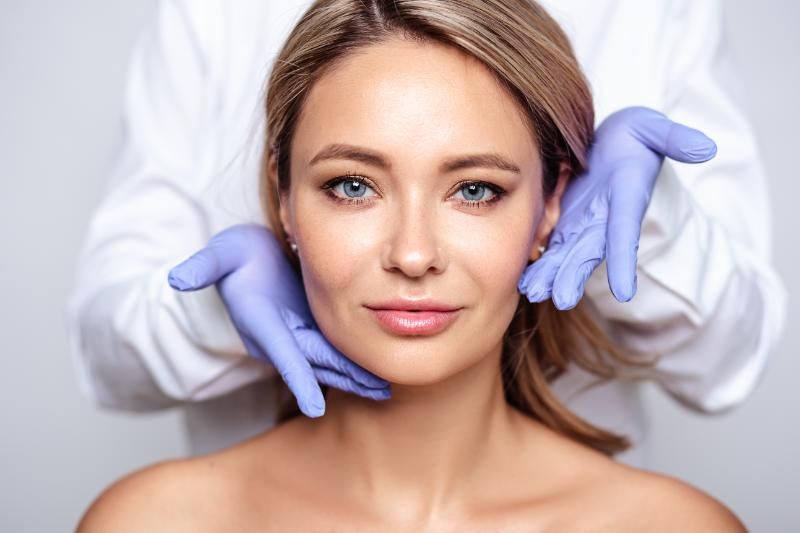 A woman faces the camera while two gloved hands rest lightly against either side of her jaw.