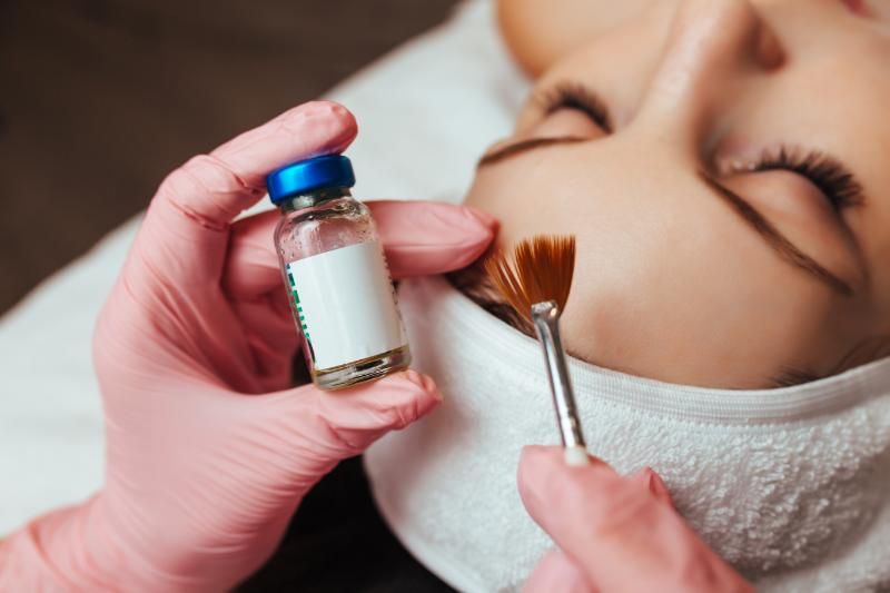A gloved hand holds a small labelled glass vial while another holds a fan brush beside the face of a woman lying with her eyes closed.