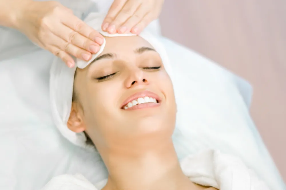 A person rests on a treatment bed in a white headwrap while another person holds a cotton pad against their forehead.