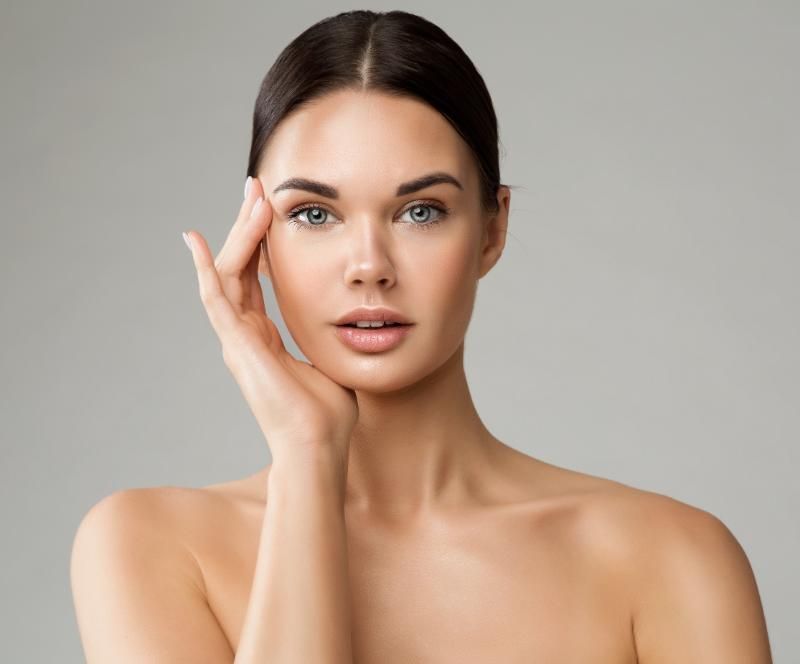 A woman with dark hair tied back raises one hand to her jaw, photographed against a plain grey background.