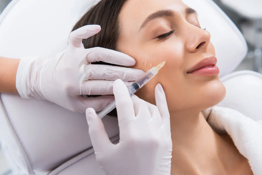 Gloved hands hold a small syringe against the cheek of a person lying back on a treatment chair with their eyes closed.