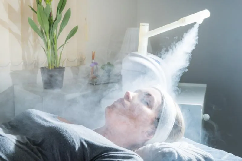 Client reclining on a treatment bed with a wrapped towel headband while a steamer directs vapour over her face