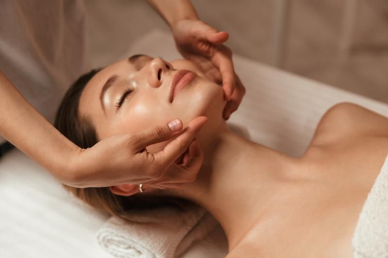 A woman lying on a treatment bed with her eyes closed while a practitioner's hands rest on her face.