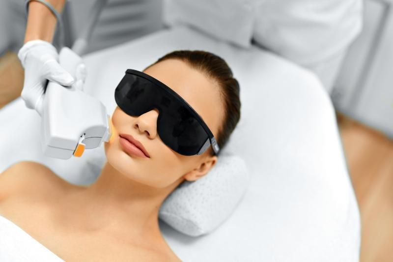 A woman reclines wearing black protective eye shields while a practitioner holds a device near her face.
