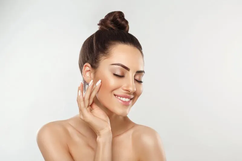 A woman with her hair in a loose bun tilts her head, eyes closed and smiling, one hand raised to her cheek.