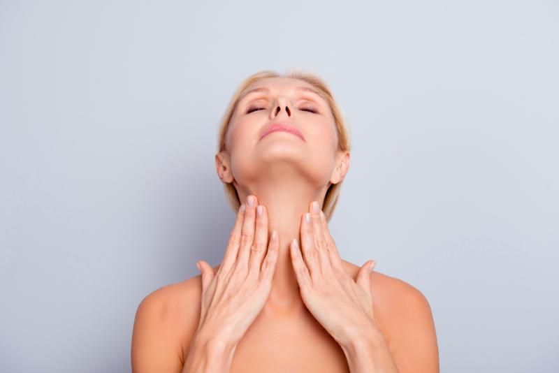 A woman tilts her head back with both hands resting on her neck, photographed against a pale blue background.