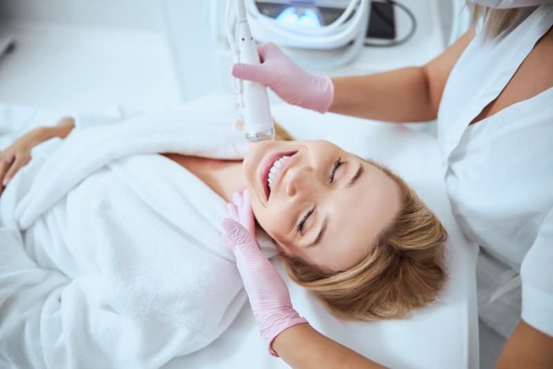 A woman wrapped in a white towel lies on a treatment bed, smiling with her eyes closed, while a gloved practitioner holds a handheld device near her jaw.