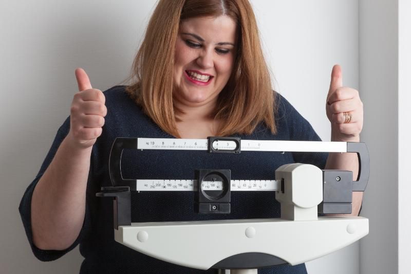 A woman standing on a medical beam scale smiles and holds up both thumbs.