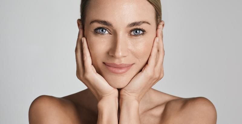 A woman looks toward the camera with a hand resting on each side of her face, against a plain grey background.